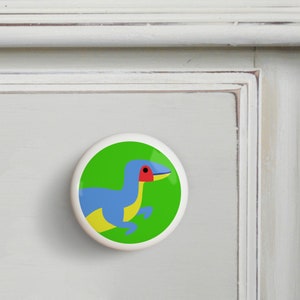 May include: A green, blue, yellow, and red dinosaur on a white ceramic knob.