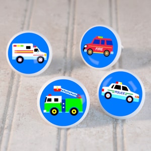 May include: Four round blue and white ceramic knobs with images of emergency vehicles. The knobs feature a red fire truck with the word "FIRE", a green fire truck with a ladder, a white ambulance, and a blue and white police car with the word "POLICE".