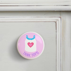 May include: Pink and white ceramic drawer knob with a cartoon tank top with a pink heart on it. The text "TANK TOPS" is written below the tank top.