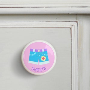 May include: Pink ceramic drawer knob with a blue and white cartoon illustration of denim shorts with a yellow flower on the pocket. The text "SHORTS" is written in purple below the shorts.