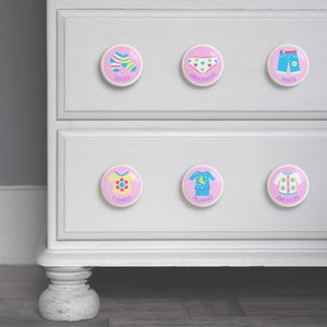May include: White dresser with six pink and white knobs. Each knob has a different clothing item on it, including socks, underwear, pants, t-shirts, pajamas, and sweaters. The knobs are labeled with the type of clothing.