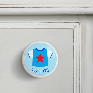 May include: A white ceramic knob with a blue and red cartoon t-shirt design. The text "T-SHIRTS" is printed below the t-shirt.