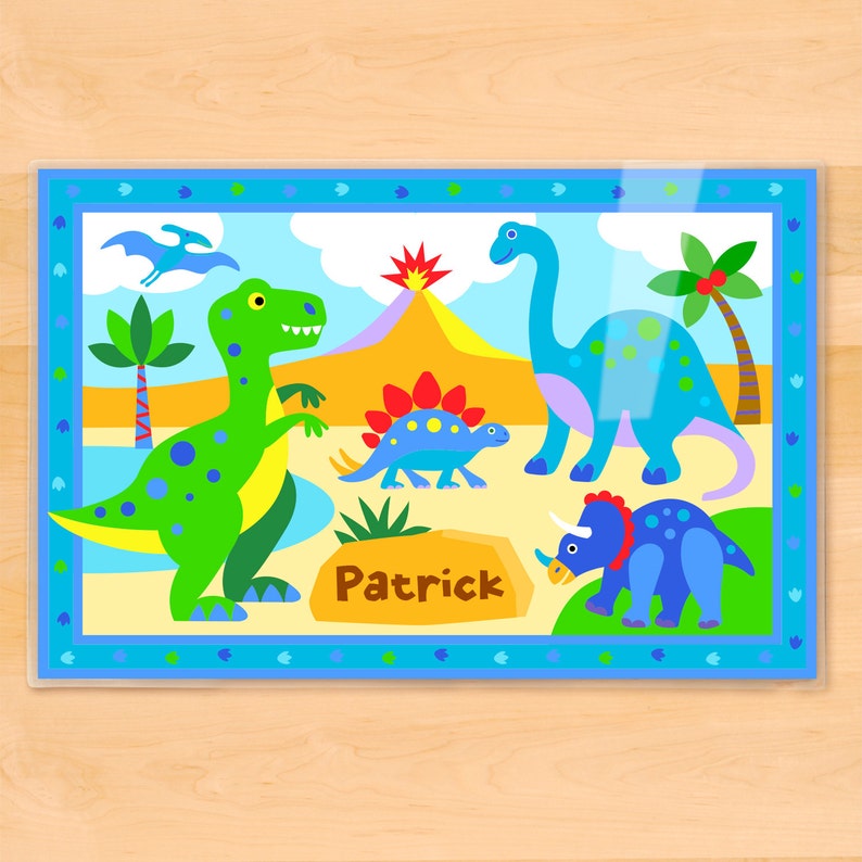 Personalized place mat with fun dinosaur illustrations. Four dinos, palm trees and a volcano in a prehistoric scene. Blue border has dino footprints. Child name in on a rock at the bottom.