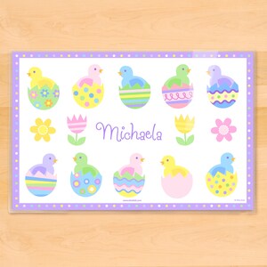 Easter place mat for kids on a wood tabletop.  Mat has pastel chicks sitting in colorful Easter eggs shells with child name in the center of the mat.  Border of mat is pastel polka dots, childs name is in soft purple.