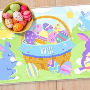 Close up of Easter place mat for kids on a wood table top with a bowl of Easter eggs. Place mat has image of an Easter basket full of eggs and personalized with name in white in the blue basket liner.