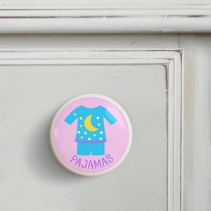 May include: A pink ceramic knob with a blue and yellow illustration of pajamas with a moon and stars. The text "PAJAMAS" is written in purple below the illustration.
