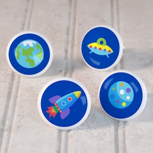 May include: Four blue and white ceramic knobs with space-themed designs. The knobs feature a rocket, a planet, a UFO, and the Earth.