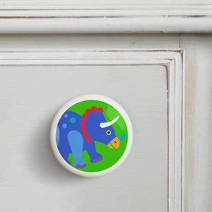 May include: A blue and green ceramic knob with a cartoon triceratops on a green background.