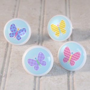 May include: Four ceramic knobs with a light blue background and colorful butterfly designs. Each knob features a different butterfly pattern with polka dots, stripes, checkered, and wavy patterns.