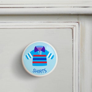 May include: A white ceramic knob with a blue background and a cartoon illustration of a striped shirt. The text "SHIRTS" is printed below the shirt.