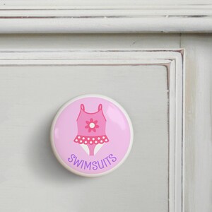 May include: Pink ceramic drawer knob with a pink and white polka dot swimsuit design and the word "SWIMSUITS" in purple text.