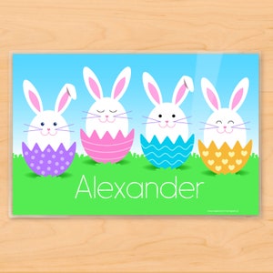 Easter Bunnies place mat for kids with 4 little bunnies, each sitting in a colorful Easter egg shell. Bunnies and eggs are on green grass with blue sky. Child's name is in white letters in grass.