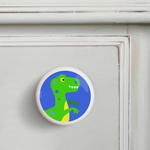 May include: A green dinosaur with yellow eyes and blue spots on a blue background. The dinosaur is facing right and has its mouth open. The dinosaur is on a white ceramic knob.