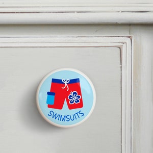 May include: A white ceramic knob with a blue background and a red and blue swimsuit design. The text "SWIMSUITS" is printed in blue below the swimsuit.