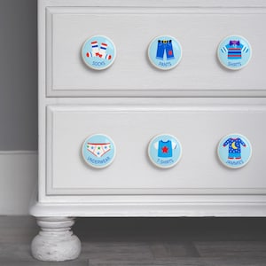 May include: White dresser with six blue and white ceramic drawer knobs. Each knob has a different image of clothing, including socks, pants, shirts, underwear, t-shirts, and jammies. The words "Socks", "Pants", "Shirts", "Underwear", "T-shirts", and "Jammies" are written below each knob.
