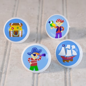 May include: Four ceramic knobs with blue backgrounds and pirate-themed illustrations. The knobs feature a treasure chest, a pirate with a parrot, a pirate with a telescope, and a pirate ship.