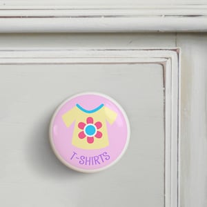 May include: Pink and yellow drawer knob with a cartoon t-shirt design and the text "T-SHIRTS" in purple.