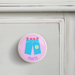 May include: A pink ceramic drawer knob with a blue cartoon pair of pants with a yellow flower on the right leg. The text "PANTS" is printed in purple below the pants.