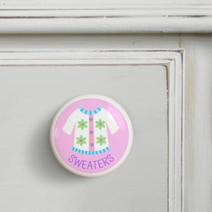 May include: Pink round ceramic drawer knob with a white sweater with green flowers and blue stripes. The text "SWEATERS" is written in purple below the sweater.