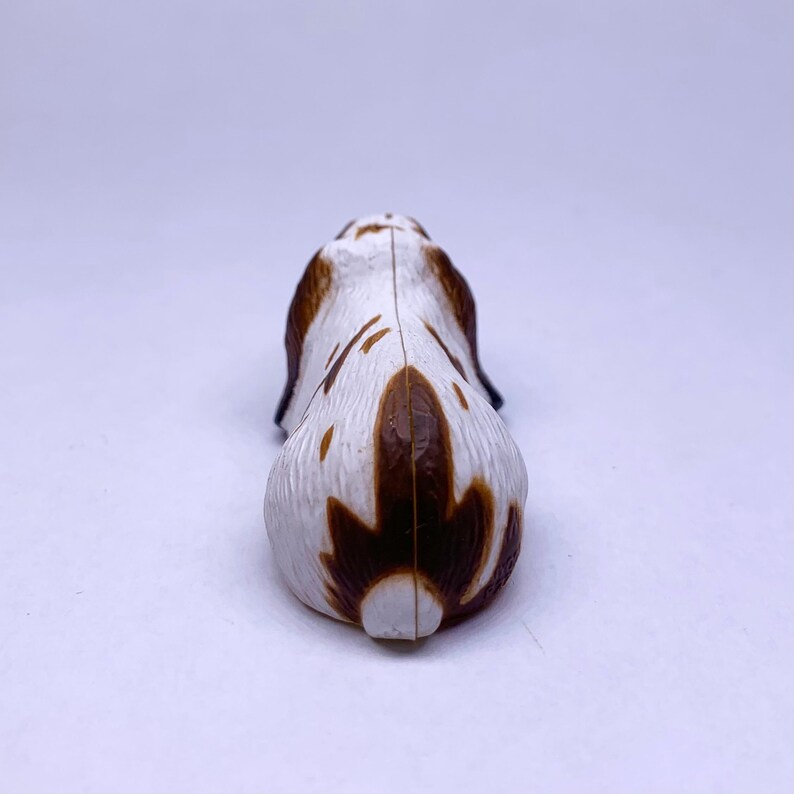 Puede incluir: Peque&ntilde;a figura de pl&aacute;stico de un perro, vista desde atr&aacute;s. El perro es principalmente blanco con manchas y marcas marrones. La figura tiene un acabado liso y brillante y est&aacute; sobre un fondo blanco.