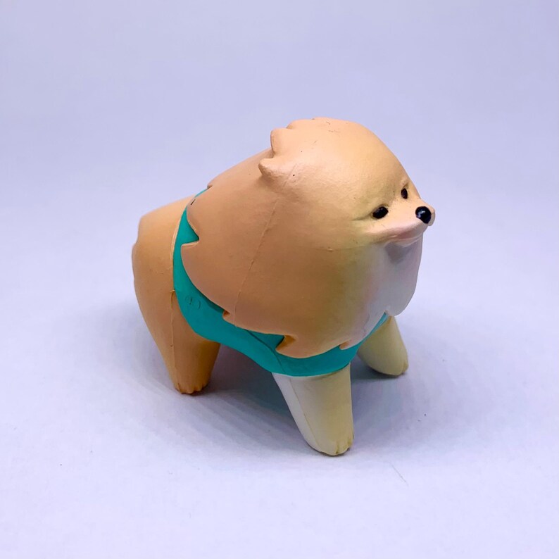 Puede incluir: Figura de perro Pomerania marr&oacute;n claro con una camisa de color turquesa. El perro tiene hocico y patas blancas, con ojos y nariz negros. La figura est&aacute; sobre una superficie blanca.