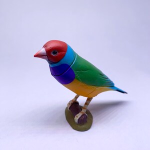 Puede incluir: Figurita colorida de un pinz&oacute;n de Gould. El ave tiene la cabeza roja, el cuello azul, el dorso verde, el vientre amarillo y la cola azul. Est&aacute; posada en una rama marr&oacute;n con una base verde. Art&iacute;culo decorativo.