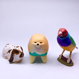 Puede incluir: Tres figuras de animales: un conejo blanco y marr&oacute;n, una figura amarilla con forma de perro con pantalones cortos turquesa y un p&aacute;jaro colorido posado en una rama. El p&aacute;jaro tiene la cabeza roja, plumas azules, verdes y amarillas.