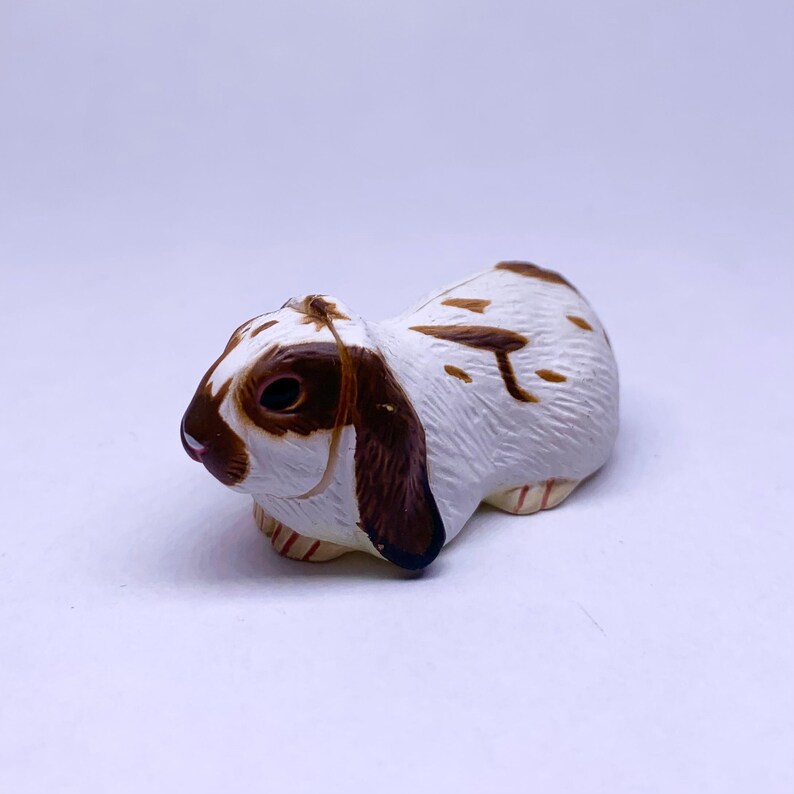 Puede incluir: Figura de conejo de cer&aacute;mica en reposo. El conejo tiene pelaje blanco con marcas marrones en la espalda, las orejas y alrededor de los ojos. La figura est&aacute; detallada con una textura realista y se encuentra sobre un fondo liso.