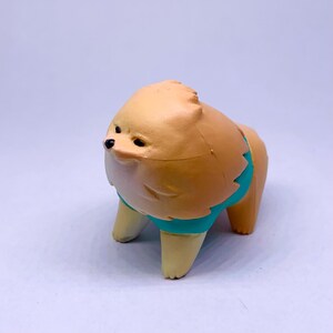 Puede incluir: Peque&ntilde;a figura de perro Pomerania de color canela con una banda turquesa alrededor de la cintura. El perro tiene ojos negros y un aspecto esponjoso, de pie sobre cuatro patas cortas. El fondo es una superficie lisa de color claro.