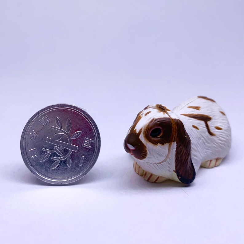 Puede incluir: Una peque&ntilde;a y detallada figura de un conejo de orejas ca&iacute;das con marcas marrones y blancas. El conejo est&aacute; colocado junto a una moneda japonesa de 1 yen, lo que proporciona una sensaci&oacute;n de escala. La figura tiene un aspecto realista.