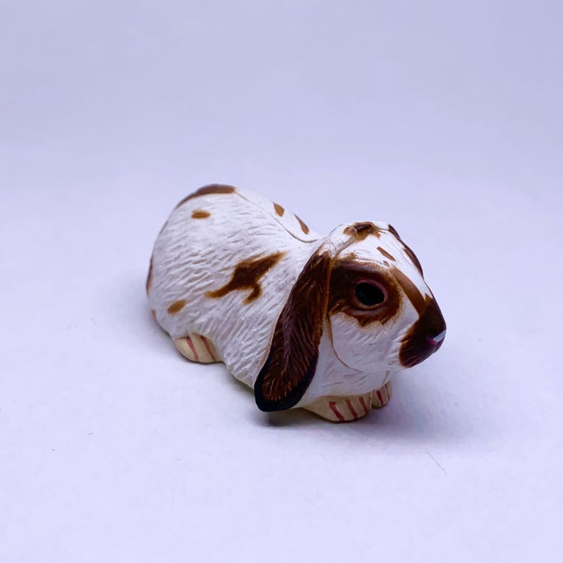 Puede incluir: Figura de conejo de cer&aacute;mica en reposo. El conejo tiene pelaje blanco con manchas y marcas marrones en las orejas y la cabeza. La figura tiene una nariz rosada y est&aacute; sobre un fondo blanco.