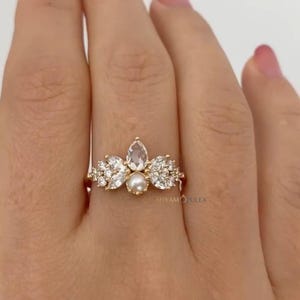 Può includere: Un anello d'oro con un gruppo di pietre preziose. L'anello presenta una pietra trasparente centrale a forma di pera, circondata da pietre trasparenti più piccole e una perla centrale. L'anello è montato su una fascia d'oro.