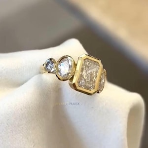 Könnte beinhalten: Ein goldener Ring mit drei Diamanten unterschiedlicher Formen und Größen. Der Ring hat einen rechteckigen Mittelstein, einen ovalen Stein und einen runden Stein. Der Ring liegt auf einer weißen Oberfläche. Der Text "SHYAM PULEX" ist sichtbar.