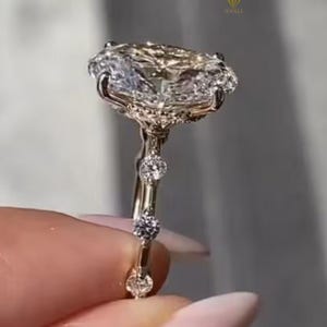 Puede incluir: Un anillo de compromiso de oro con un gran diamante de talla ovalada como pieza central. El anillo presenta diamantes más pequeños engastados a lo largo de la banda, añadiendo brillo. El anillo se sostiene sobre un fondo borroso.