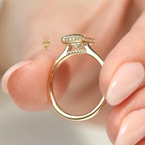 May include: A gold ring with a square-cut, clear gemstone and small accent stones. The ring is held between fingers, showcasing the intricate design and sparkle of the jewelry. The band is a simple, polished gold.
