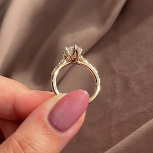 May include: A gold engagement ring with a large, clear gemstone. The ring features a delicate band with small accent stones and a detailed setting that holds the main stone. The ring is held up against a soft, brown background.