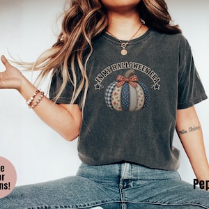 May include: A charcoal gray short-sleeve t-shirt featuring a graphic of a patchwork pumpkin with a plaid bow. The text above the pumpkin reads IN MY HALLOWEEN ERA, flanked by two small stars. The shirt has a crew neckline and a relaxed fit.
