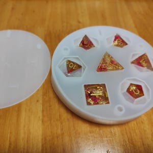 May include: A white silicone mold with a lid, containing several resin dice in various shapes. The dice are transparent with red and gold accents, and the numbers are gold. The mold is on a wooden surface.