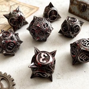 Puede incluir: Un juego de siete dados metálicos de varias caras con un acabado en color gris plomo oscuro, puntas afiladas y detalles en pintura roja. Cada dado tiene números en relieve de tono plateado y una superficie texturizada con patrones geométricos.