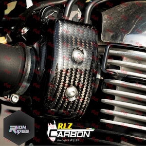 May include: A carbon fibre motorcycle component with a woven design, secured with two silver bolts. The part is black and grey, and is mounted on a motorcycle engine. The image also includes the text "RLZ CARBON" and "ION RIDES".