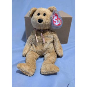 Könnte beinhalten: Ein hellbrauner Plüschteddybär mit schwarzer Nase und kleinen schwarzen Augen. Der Bär hat ein rosa Band um den Hals und ein rot-weißes Etikett an seinem Ohr. Der Bär sitzt vor einem Pappkarton.