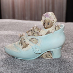 May include: A blue ceramic shoe figurine with a mother mouse and her babies inside. The mother mouse is wearing a pink and white hat. The shoe has a white strap and a white heel.