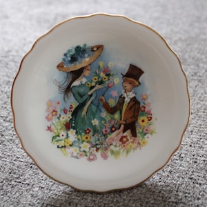 May include: A white porcelain trinket dish with a gold rim features a painted scene of two figures in a garden setting. The woman wears a blue hat and a green dress, while the man wears a brown top hat and a brown jacket. The scene is surrounded by colorful flowers.