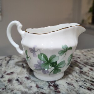 May include: A white ceramic creamer with a gold rim and a handle. The creamer is decorated with green and purple shamrock designs. The creamer is sitting on a marble countertop.