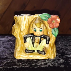 May include: A vintage ceramic planter shaped like a tree trunk, featuring a seated figure with green eyes, wearing a black jacket and yellow pants. A red flower and green leaves adorn the side. The planter is painted in shades of yellow, brown, and black.