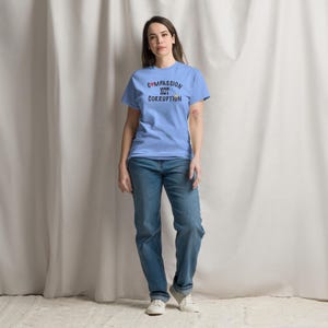 May include: A light blue t-shirt with the words "COMPASSION NOT CORRUPTION" printed in black, red, and yellow. The person is wearing blue jeans and white sneakers. The background is a white curtain.