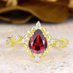 Pode incluir: Um anel de ouro com uma pedra preciosa granada vermelha em forma de pera no centro, rodeada por uma moldura decorativa de pequenas pedras claras. A banda tem um design texturizado de videira com pedras claras adicionais nas laterais.