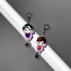 May include: Two anime-style keychains featuring male characters in suits. One keychain has a character with black and white hair, the other with brown hair. Both have star-shaped clasps and small, decorative charms.