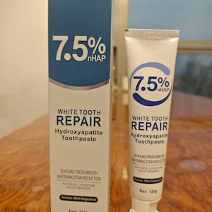 May include: A white tube and box of toothpaste with blue accents. The text on the packaging reads "7.5% nHAP WHITE TOOTH REPAIR Hydroxyapatite Toothpaste". The tube and box are on a wooden surface. The net weight is 120g.