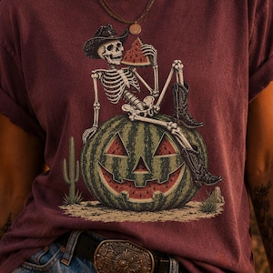 Op de afbeelding: Een donkerrood t-shirt met een afbeelding van een skelet met een cowboyhoed en laarzen, zittend op een grote watermeloen die is uitgesneden als een pompoenlantaarn. Het skelet houdt een stuk watermeloen vast, met een kleine cactus ernaast.